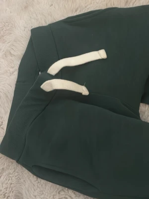 Mörkgröna mjukisbyxor Gina Tricot XS - Mörkgröna mjukisbyxor från Gina Tricot i storlek XS. Byxorna har bred resår i midjan, vita snören för justering och en bakficka med broderad detalj. Perfekta för chill dagar eller när du vill vara extra bekväm.