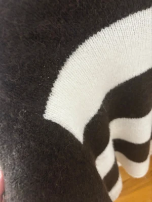 Randig brunvit stickad tröja - Säljer en mysig stickad tröja med breda svarta och vita ränder. Tröjan har lång ärm och en avslappnad passform, perfekt för kyliga dagar. Materialet känns mjukt och fluffigt, vilket gör den extra skön att bära.