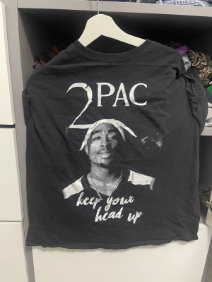 Svart 2Pac t-shirt med tryck - Svart t-shirt med stort 2Pac-tryck på ryggen och texten 'keep your head up' i vitt. Klassisk passform med korta ärmar och rund hals. Perfekt för dig som gillar streetstyle och hiphop-vibe.