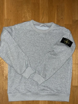 Stone island grå - Säljer en stilren grå sweatshirt från Stone Island med klassisk rund hals och ribbade muddar. Tröjan har den ikoniska Stone Island-loggan på ärmen och är gjord i mjuk bomull för maximal komfort.