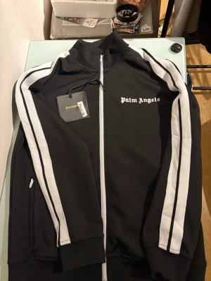 Palm Angels svart track jacket - Svart track jacket från Palm Angels med vita ränder längs ärmarna och vit dragkedja. Klassisk hög krage och logga på bröstet. Sportig vibe och perfekt för streetwear. Materialet är polyester och modellen har långa ärmar med muddar.