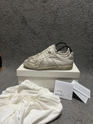 Maison Margiela - Maison Margiela sneakers i vit canvas med distressed och sliten look. Skorna har snörning, rund tå och platt sula. Unik design med råa kanter och vintage-inspirerade detaljer. Kommer med originalbox och dustbag.