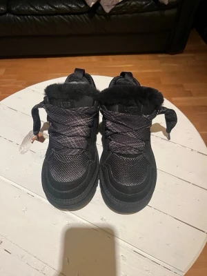 Ugg lowmel  - Säljer ett par svarta UGG sneakers med breda snören och fluffig fuskpäls runt öppningen. Skorna har en mix av mocka och textil på ovandelen samt en platt sula. Perfekta för dig som vill ha både värme och stil under höst och vinter.