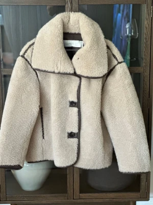 Beige teddyjacka från ARKET XS - Säljer en beige teddyjacka från ARKET i storlek XS. Jackan har stora kragar, markerade mörkbruna sömmar och knäppning med knappar framtill. Utsidan är fluffig och mjuk, perfekt för kyliga dagar. Snygg och trendig modell som ger en cool look.