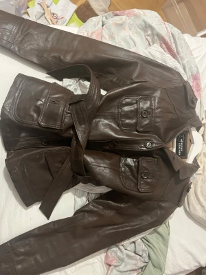 Brun skinnjacka med bälte - Säljer en brun skinnjacka från Genuine Leather med fyra stora fickor framtill, knappar hela vägen och ett matchande bälte i midjan. Jackan har klassisk krage och axelklaffar för en cool look. Perfekt för dig som vill ha en tidlös och snygg jacka.