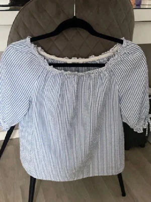 Blårandig offshoulder blus - Söt blus med blå och vita ränder, offshoulder-modell och puffiga korta ärmar med knyt i ärmsluten. Blusen har en luftig och avslappnad passform, perfekt för varma dagar. Tillverkad i ett lätt bomullstyg.