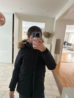 Svart parkasjacka med fuskpäls - Säljer en svart parkasjacka från Mountain Works med mysig vit teddyfoder på insidan och huva med fuskpäls. Jackan har dragkedja och knappar framtill samt två stora fickor. Perfekt för kalla dagar och riktigt skön att bära.