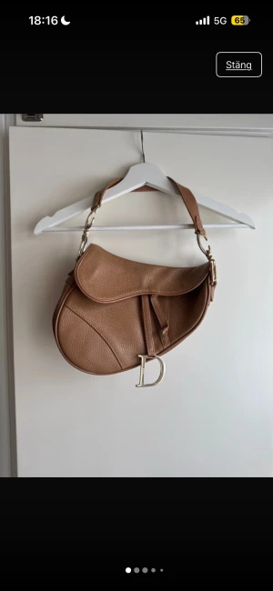 Brun Saddle Bag  - Brun sadelväska köpt second hand. Jag är osäker på både material och äkthet, därför säljs väskan som icke verifierad.  Väskan är i bruksskick och har: – Slitage på metalldelarna där färgen har nötts - Slitningar på färgen på remmen – Ena remmen är sliten men sitter fortfarande fast och fungerar se bild  Lite smutsig inuti  – Mindre skavanker förekommer (se bilder)  Materialet kan vara läder eller konstläder, jag kan tyvärr inte garantera vilket. Passar bra som vardagsväska eller för dig som vill