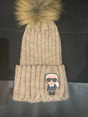 Beige stickad mössa med pälsboll - Snygg beige stickad mössa med stor pälsboll på toppen. Mössan har ribbad struktur och en cool patch framtill med en tecknad figur. Perfekt accessoar för att hålla värmen och se trendig ut under kalla dagar.