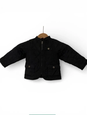 Svart quiltad jacka från Gant - Quiltad svart jacka från Gant med dragkedja framtill och två fickor med knapp. Jackan har en klassisk krage och diskret röd logga broderad i nacken. Insidan har rutigt foder upptill och jackan är perfekt för kyliga dagar.