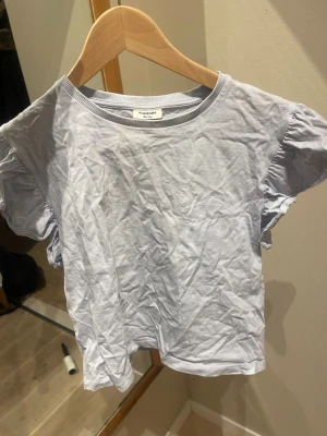 Ljusblå volangärmad topp Kappahl - Säljer en ljusblå topp från Kappahl i storlek 158/164. Toppen har korta volangärmar och rund halsringning med ribbad kant. Tillverkad i mjuk bomull, perfekt för en enkel och fräsch look. Kan även strykas inna jag skickar 