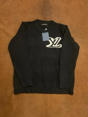 Svart stickad tröja från Louis Vuitton - Svart stickad tröja från Louis Vuitton med stor vit LV-logga på bröstet. Tröjan har rund halsringning, ribbade muddar och är långärmad. Perfekt för dig som vill ha en clean och lyxig look med tydlig designerstil.