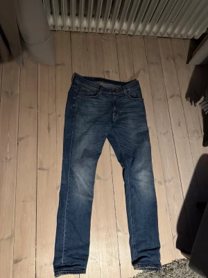 Blåa nudie jeans  - Säljer ett par klassiska blå jeans med raka ben och snygg tvätt. Jeansen har fem fickor, normal passform och subtila slitningar framtill. Bakfickorna har dekorativa sömmar och det finns en läderpatch i midjan. Modellen är ”lean dean” Perfekta till vardagslooken.