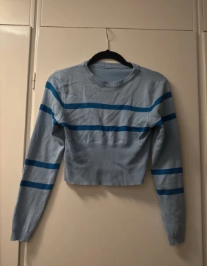 Ljusblå randig croppad tröja - Säljer en ljusblå croppad tröja med mörkblå horisontella ränder. Tröjan har rund halsringning, långa ärmar och ribbade muddar. Perfekt för dig som gillar en chill och trendig stil.