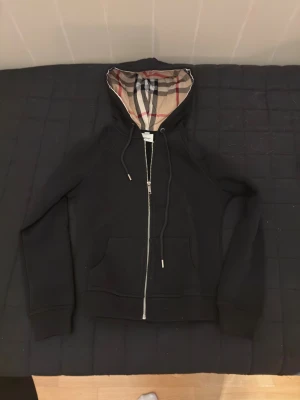 Svart Burberry hoodie med dragkedja - Svart hoodie från Burberry med klassiskt rutigt foder i huvan. Tröjan har dragkedja framtill, snörning vid huvan och två fickor. Långärmad och tillverkad i mjukt bomullsmaterial. Knappast använd och ganska liten i storlek.  Säljer då den inte passar mig längre. 