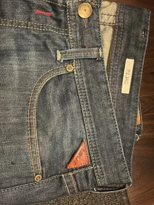 Replay blå jeans med slitningar - Säljer ett par blå Replay jeans med snygga slitningar och kontrastsömmar. Klassisk femficksmodell med detaljer i läder och metallknappar. Jeansen har en rak passform och tydliga slitningar framtill och baktill för en cool, använd look. Storlek 34/32 men passar mig bra som har 30/32 och 31/32
