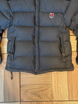 Mörkblå pufferjacka från Fjällräven - Säljer en mörkblå pufferjacka från Fjällräven med klassisk logga på bröstet. Jackan har två dragkedjeförsedda fickor framtill, quiltad design och justerbar nederkant med snörning. Perfekt för kalla dagar och riktigt snygg till streetwear.