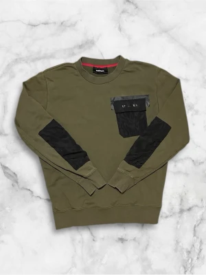 Olivgrön sweatshirt från Diesel - Säljer en olivgrön sweatshirt från Diesel med svarta detaljer på armbågarna och en stor svart ficka med lock på bröstet. Tröjan har rund halsringning och ribbade muddar. Perfekt för dig som gillar streetwear och vill ha något unikt i garderoben.