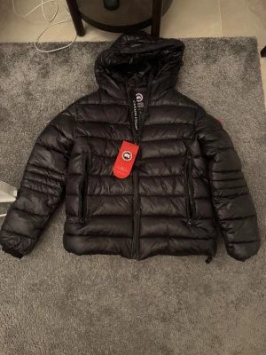 Svart dunjacka från Canada Goose S - Säljer en svart dunjacka från Canada Goose med huva och dragkedja framtill. Jackan har två stora fickor med dragkedja och den klassiska Canada Goose-loggan på ärmen. Perfekt för kalla dagar och riktigt snygg passform.