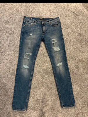 Jack & Jones skinny fit jeans blå - Säljer ett par blå skinny fit jeans från Jack & Jones, modell Liam. Jeansen har slitna detaljer och hål på både fram- och baksida, klassisk femficksdesign och orangea sömmar. Perfekta för dig som gillar en smal passform och lite ruffig look. W32 L30 Passar mer som w30