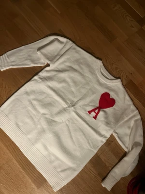 Vit stickad tröja från Ami Paris - Säljer en vit stickad tröja från Ami Paris med röd hjärtdetalj och bokstaven A på bröstet. Tröjan har rund halsringning, långa ärmar och ribbade muddar vid ärmslut och nederkant. Perfekt för dig som gillar stilrena plagg med en twist.