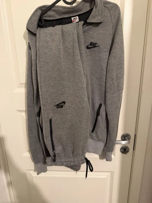 Grå träningsväst från Nike - Säljer en grå träningsväst från Nike med svart broderad logga och texten 'NIKE RUNNING' på axeln. Västen har dragkedja framtill, två fickor med dragkedja och justerbar nederkant med snörning. Materialet är mjukt och andas, perfekt för löprundan.