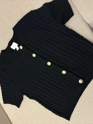 Svart kabelstickad kofta från H&M - Svart kabelstickad kofta från H&M med korta ärmar och guldiga knappar på framsidan. Koftan är köpt 2024 och har använts sparsamt, den var viral ett tag och är verkligen så fin🥰Den är storlek L. 