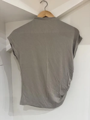 Beige topp med rynkade detaljer 157 - Säljer en beige topp från 157 i storlek M. Toppen har korta ärmar och en snygg rynkad detalj på ena sidan som ger en cool twist. Tillverkad i mjuk bomull och har en enkel, stilren look som passar till mycket.