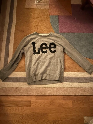 Grå sweatshirt från Lee - Säljer en grå sweatshirt från Lee med stort svart tryck på bröstet. Tröjan har rund halsringning, långa ärmar och ribbade muddar vid ärmslut och nederkant. Perfekt för en chill och avslappnad stil.