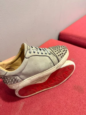 Gråa Christian Louboutin sneakers med nitar - Säljer ett par gråa sneakers från Christian Louboutin strl 42 med ikoniska röda sulor och coola nitar på tå och häl. Skorna har snörning och är tillverkade i mocka med läderfoder. Perfekt för dig som vill sticka ut med en edgy och lyxig stil.