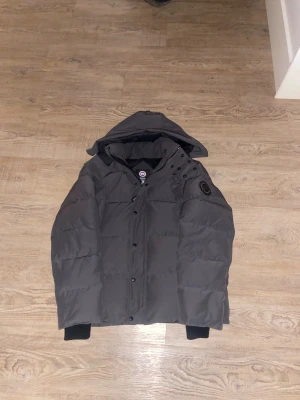 Svart pufferjacka från Canada Goose - Säljer en svart pufferjacka från Canada Goose med huva och snygga detaljer vid axeln. Jackan har broderad logga på ärmen, dragkedja och knappar framtill samt ribbade muddar vid ärmslut. Perfekt för kalla dagar och har en clean, modern look. den är använd 4 gånger så inprincip ny 