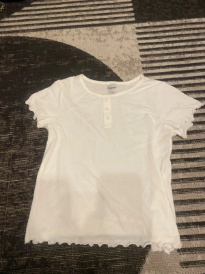 Vit ribbad topp från Kappahl - Säljer en vit ribbad topp från Kappahl med korta ärmar och rund hals. Toppen har dekorativa knappar framtill och vågiga kanter vid ärmslut och nederkant som ger en söt touch. Perfekt att styla med jeans eller kjol för en fräsch look.