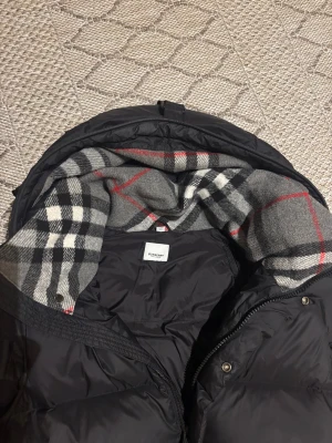 Svart Burberry pufferjacka med huva - Svart pufferjacka från Burberry med huva och klassiskt rutigt foder i grått, vitt och rött. Jackan har tre fickor, där en av de på insidan. Perfekt för kalla dagar och riktigt snygg streetstil.