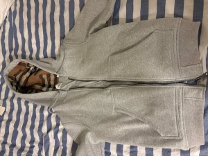 Grå hoodie från Burberry med dragkedja - Säljer en grå hoodie från Burberry med klassiskt rutigt foder i huvan. Tröjan har två stora fickor framtill, ribbade muddar och en snygg broderad logga på bröstet. Dragkedja hela vägen och mjukt bomullsmaterial. Perfekt för dig som gillar stilren design med lyxig touch.