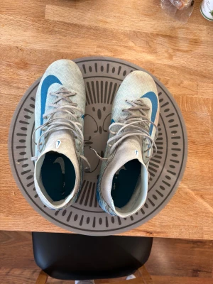 Nike Mercurial blå fotbollsskor - Säljer ett par Nike Mercurial fotbollsskor i ljusgblått med mörkblåa detaljer och stor Nike-logga på sidan. Skorna har snörning, högre skaft och platta dobbar för bra grepp på planen. Perfekta för dig som vill sticka ut på matchen!