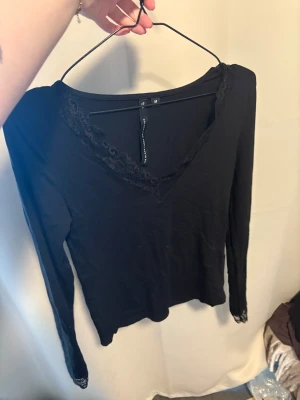 Svart långärmad topp med spets - Svart långärmad topp med v-ringning och fina spetsdetaljer runt halsen och vid ärmsluten. Tunn och mjuk i materialet, perfekt för att styla med jeans eller kjol. Enkel men ändå elegant look.