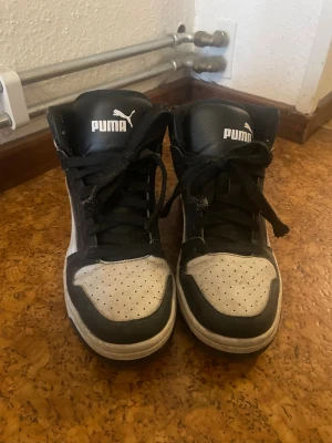 Svarta och vita Puma high-top sneakers - Säljer ett par high-top sneakers från Puma i svart och vitt. Skorna har snörning, perforerad tå och klassisk Puma-logga på sidan och plösen. Materialet är syntet och textil, med platt sula och rund tå. Perfekt för dig som gillar sportig streetstil.