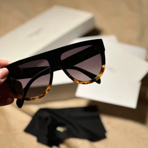 Svarta Celine solglasögon - Snygga solglasögon från Celine med bred svart båge och detaljer i sköldpaddsmönster längst ner. Glasen är mörka och har en lätt toning.  Haft dem ett tag så det finns tecken på användning, men inget som är synligt när man har på sig dem. Nypris var ca 3600 om jag minns rätt.