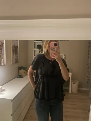 Grå blus med broderi och knyt - Supersöt svart blus från Odd Molly med broderade blommor på ryggen. Blusen har korta ärmar, knyt i halsen, volangdetaljer och spetskant nertill. Luftig passform och mjukt bomullsmaterial, perfekt till jeans eller kjol.