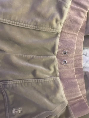 Beige mjukisbyxor Juicy Couture S - Snygga beige mjukisbyxor från Juicy Couture i storlek S. Byxorna har bred resår i midjan med metallöljetter, två stora fickor fram och bak med knappdetaljer. Mjukt velourmaterial och straight fit för chill vibe.