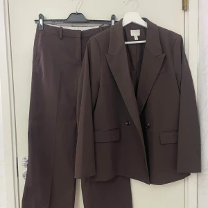 Brun kostymset från H&M - Säljer ett brunt kostymset från H&M med klassisk kavaj och raka kostymbyxor. Kavajen har en oversized look, dubbla knappar och fickor med lock. Byxorna har pressveck, bälteshällor och bakfickor med lock. Perfekt för en stilren look.