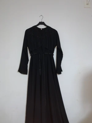 Svart långklänning med snörning - Svart långklänning med elegant snörning i midjan och subtil glans på överdelen. Klänningen har långa ärmar med volangdetaljer vid ärmsluten och ett flytande fall. Materialet känns mjukt och har en lätt struktur, perfekt för dig som gillar en klassisk och stilren look.