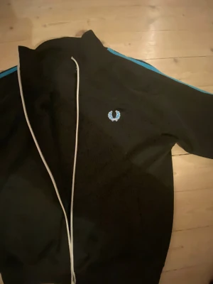Svart Fred Perry vindjacka med blå detaljer - Svart jacka från Fred Perry med blå ränder längs axlarna och vitt broderat logomärke på bröstet. Jackan har ribbad krage, dragkedja framtill och är långärmad. Materialet är troligtvis polyester och jackan har en sportig, normal passform. Lite färgskav på dragkedjan är enda tecknet på användning.
