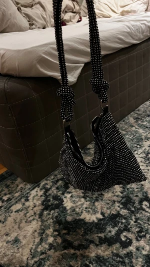 Glittrig svart axelväska med strass - Säljer en svart axelväska täckt med glittrande strass och snygga knutdetaljer på axelremmen. Väskan har en dragkedja upptill och insidan är fodrad i svart tyg. Perfekt för att lyfta din outfit med lite extra bling.