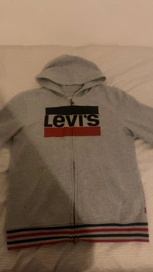 Levis hoodie  - Snygg grå hoodie från Levi's med stor logga i svart och rött på bröstet. Tröjan har huva, dragkedja framtill och ribbade muddar med röd, blå och vit rand längst ner. Perfekt för chill dagar och streetstyle.