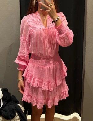Rosa volangklänning med v-ringning - Supersöt kort rosa klänning med v-ringad hals och långa ärmar. Klänningen har flera lager med volanger och smock i midjan för en snygg passform. Materialet känns lätt och luftigt, perfekt för varmare dagar. Slut såld svår att hitta.