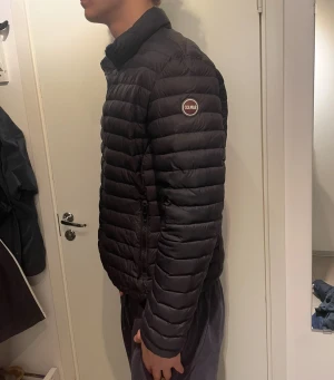 Colmar jacka - Svart pufferjacka från Colmar med diskret logga på ärmen. Jackan har horisontella quiltade paneler, dragkedja framtill och ståkrage. Materialet är lätt och syntetiskt, perfekt för kyliga dagar. Snygg och enkel design som funkar till det mesta. Storlek 56 står det på lappen, sitter som L.