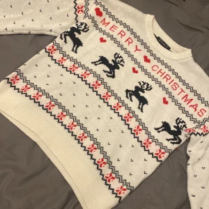 Jultröja med renar från Simplee - Vit stickad jultröja från Simplee med svart och rött mönster. Tröjan har motiv av renar, hjärtan och texten 'Merry Christmas' över bröstet. Rund hals och långa ärmar, perfekt för att maxa julstämningen. Mönstrad med klassiska juldetaljer och ribbade muddar.