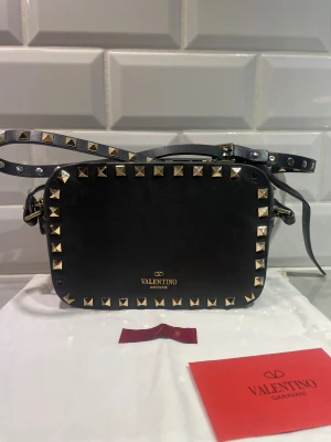 Valentino rockstud väska  - Säljer min favvis väska!! Dessvärre har 2 nitar på framsidan ramlat av, bortsett från det är skicket utmärkt 💖💖 Väskan är svart med guldiga nitar och detaljer 🥰  Väskan är köpt begagnat vilket gör att jag inte kan garantera dess äkthet, fick med en dustbag och själva kuvertet där äkthetsbeviset ligger men dessvärre inte själva korten, därav lägre pris 🥹🥹💖
