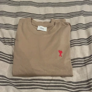 Beige t-shirt från Ami med rött hjärta - Snygg beige t-shirt från Ami Alexandre Mattiussi med klassisk passform och korta ärmar. På bröstet finns ett broderat rött hjärta med bokstaven A. Tillverkad i mjuk bomull som känns skön mot huden. Perfekt för dig som gillar stilrena detaljer. Passar även storlek L. Nypris ligger runt 2000kr